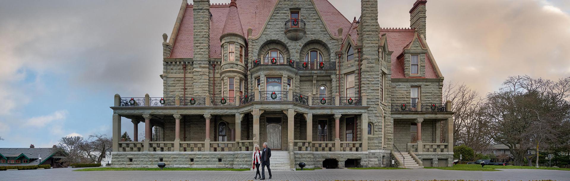 A couple walks in front of the the facade of Craigdarroch Castle Historic House & Museum adorned with holiday wreaths in Victoria, BC.