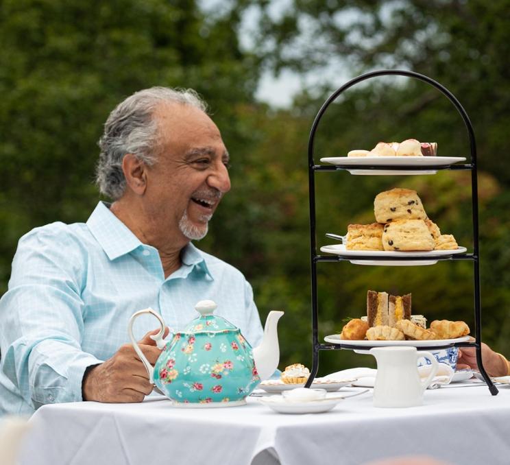A man in a blue shirt enjoys an afternoon tea experience at a table set with a blue tea pot and three-tier tower of baked goods at Abkhazi Garden in Victoria, BC