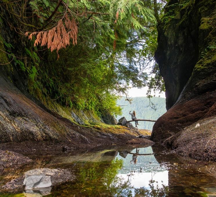 A woman sits on a log across a small cove at Wild Renfrew in Port Renfrew just outside of Victoria, BC