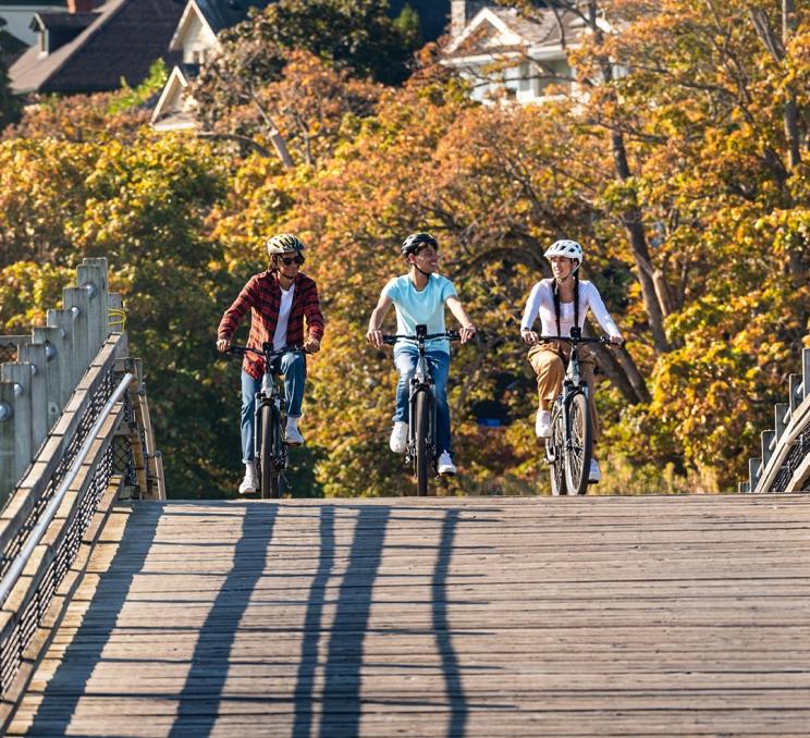 Three friends ride bikes over the Selkirk Trestle along the Galloping Goose Regional Trail, backdropped by yellow fall foliage, in Victoria, BC