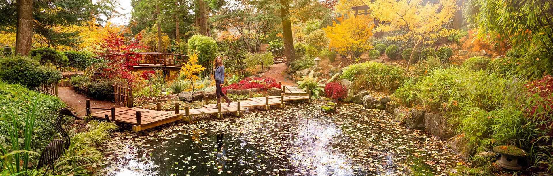 A woman in a denim coat walks across a bridge in a gardens during fall in Victoria, BC