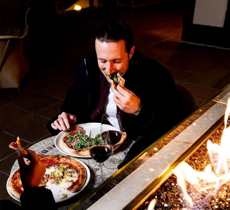A man bites into a slice of pizza next to an outdoor fireplace on the patio during with winter at FARO Handcrafted Pizza and Tasting Room in Victoria, BC