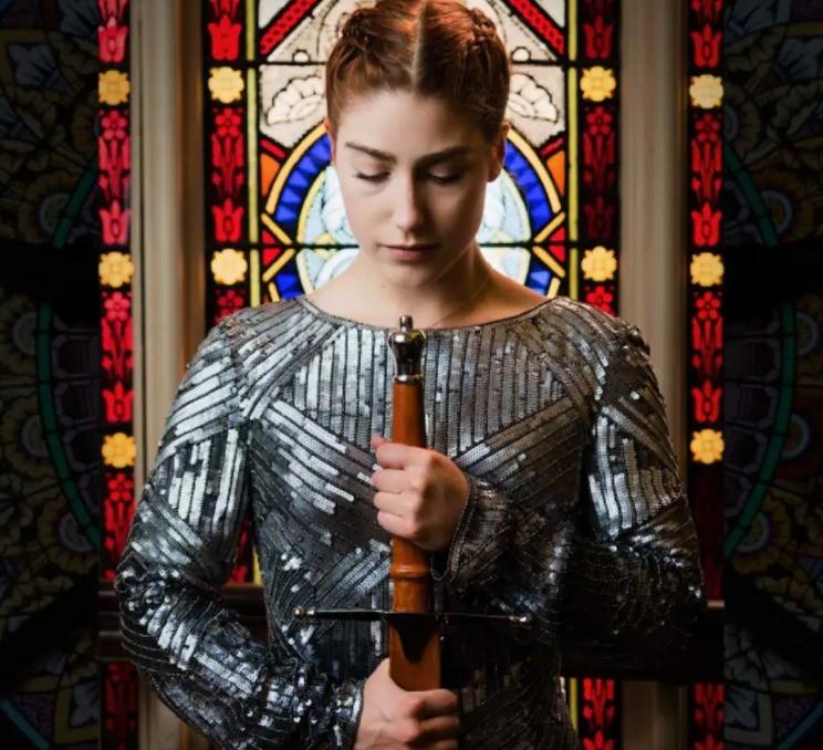A performer dressed in metal armor holding a sword stands in front of a stained glass window during a performance of JÉHANNE in Victoria, BC