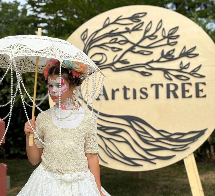 A woman in a white Victorian era dress and face paint holds a white sun umbrella at the ArtisTREE Festival at Government House in Victoria, BC