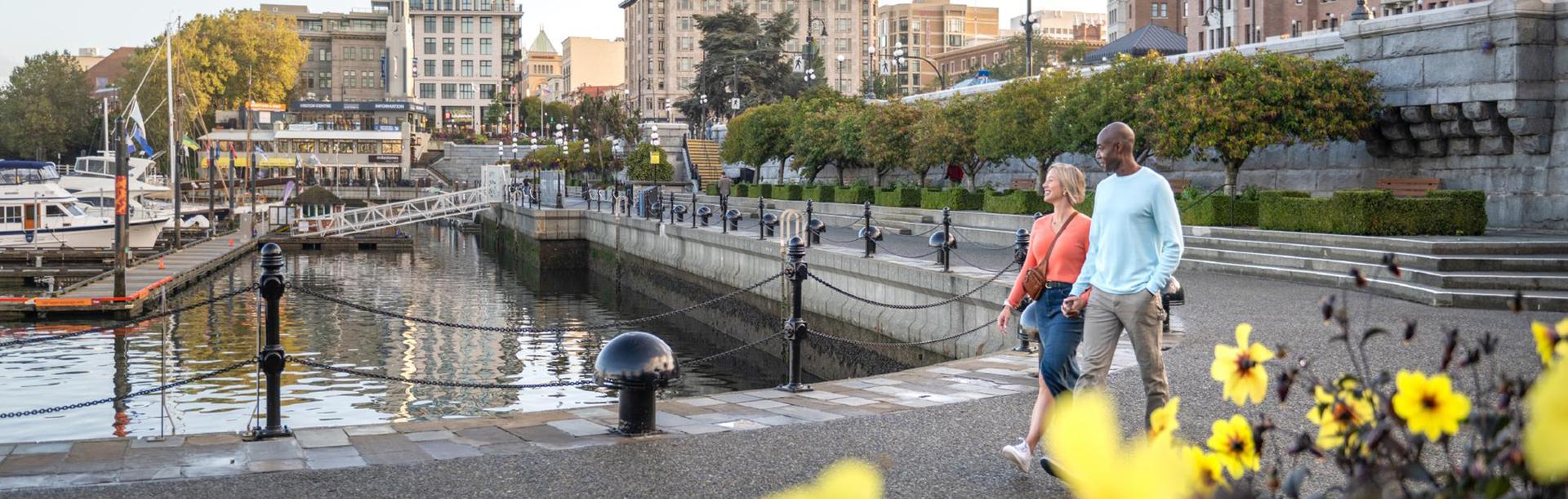 A woman wearing a pink top and a man wearing a blue sweater walk along the lower causeway of the Inner Harbour in Victoria, BC