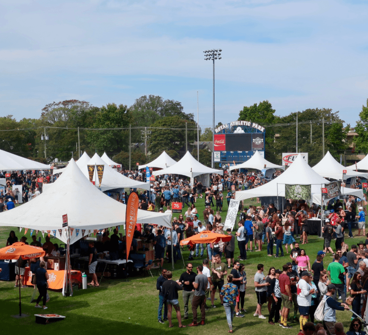 Crowds gather among white tents at the Great Canadian Beer Festival in Royal Athletic Park, enjoying craft beer tastings on a sunny day in Victoria, BC.