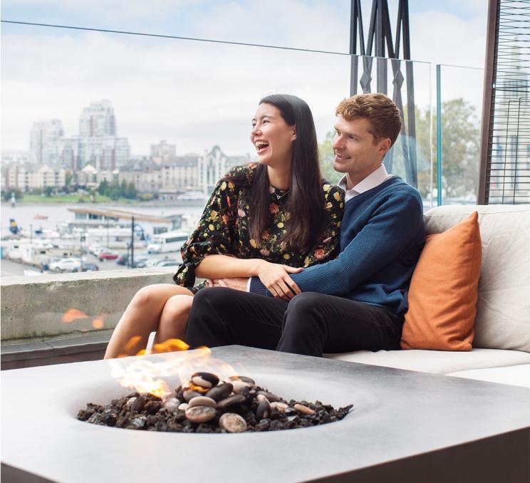 A young couple sit behind a fire, on patio furniture with an orange pillow on the veranda at the Fairmont Empress overlooking Victoria's Inner Harbour