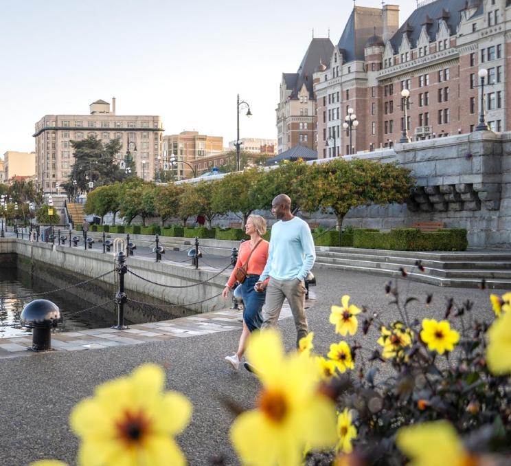 A woman in a pink top and a man in a blue sweater walk behind a flower bed filled with yellow flowers on the lower causeway of the Inner Harbour in Victoria, BC