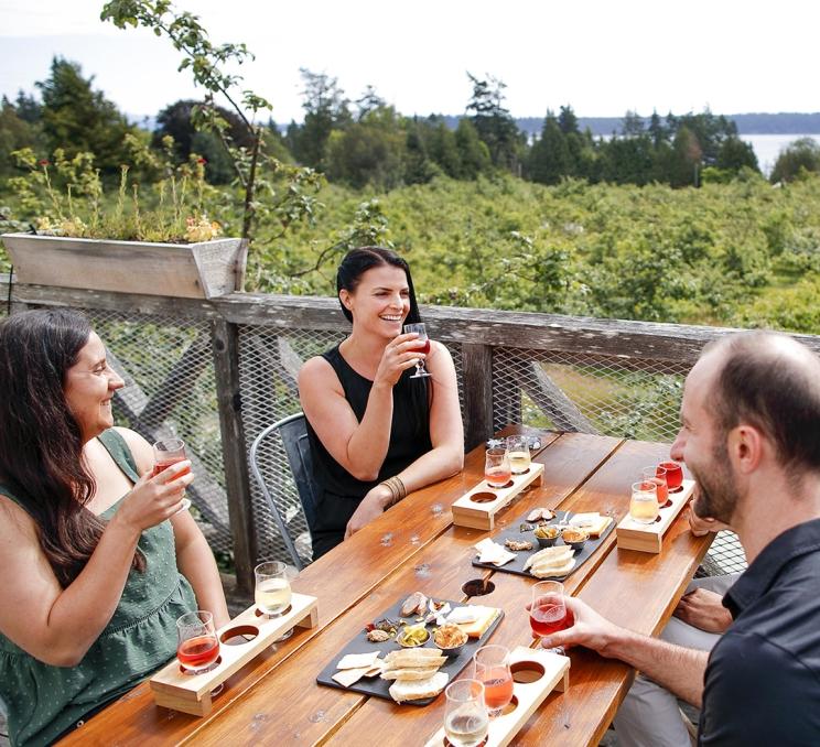 A group of friends enjoy cider flights and charcuterie while dining on the patio and overlooking the orchard at Sea Cider Farm & Ciderhouse in Victoria, BC