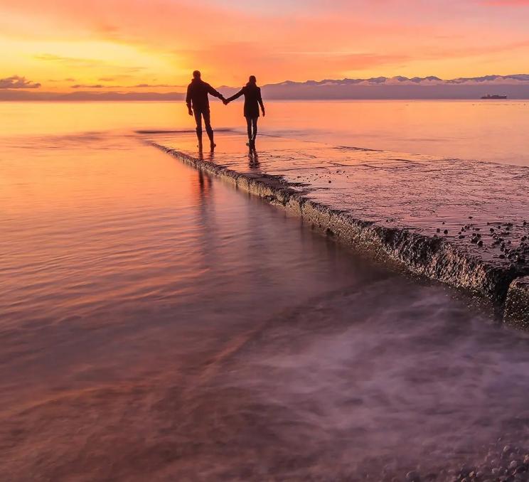 A couple walks hand in hand down a pathway to the sea in Victoria, BC at sunset
