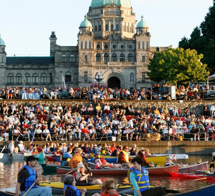 A crowd, including kayakers on the water, gathers outside the BC Parliament Buildings along the Inner Harbour for Symphony Splash! in Victoria, BC