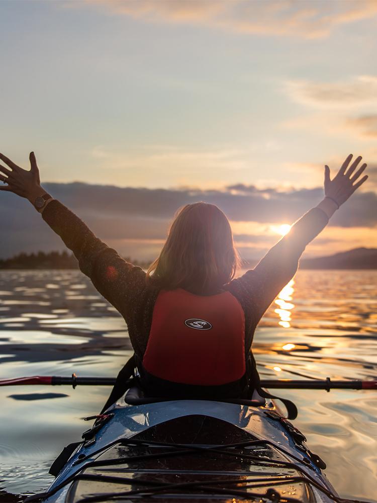 A woman in a red life preserver waves her arms in the air while enjoying a sunset while kayaking in the Salish Sea in Victoria, BC