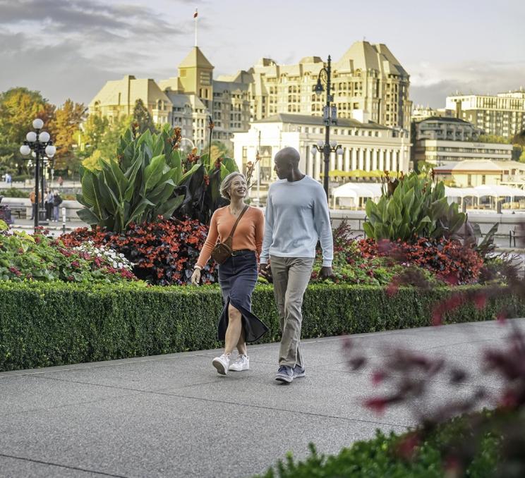 A woman wearing pink and a man in a blue sweater walk up the walkway through the gardens at the Fairmont Empress hotel in Victoria, BC