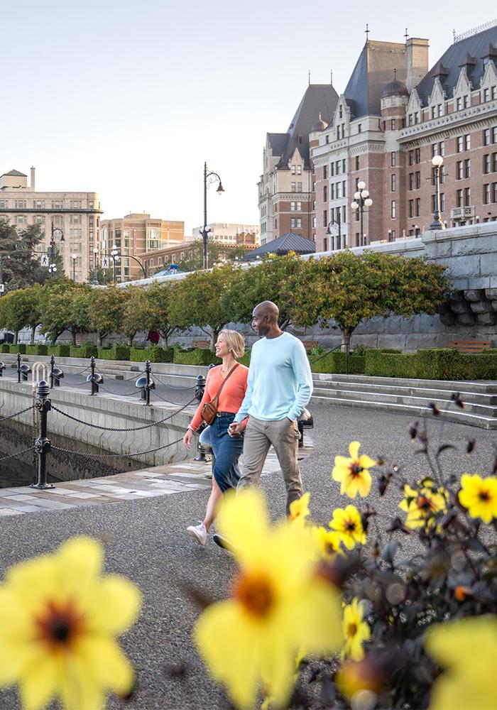 A woman in a pink sweater walks hand-in-hand with a man in blue sweater behind some yellow flowers along the Lower Causeway of the Inner Harbour with the Fairmont Empress in the background in Victoria, BC
