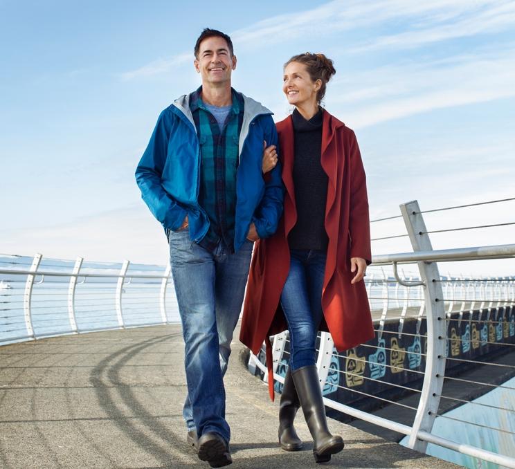 A man in a blue jacket and a woman in a red coat walk, arms linked, down the Ogden Point Breakwater in Victoria, BC