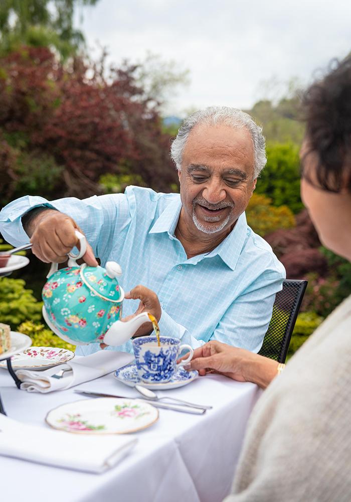 A man in a blue shirt pours tea out of a turquoise teapot for his wife while sat on the patio at Abkhazi Teahouse in Abkhazi Garden in Victoria, BC