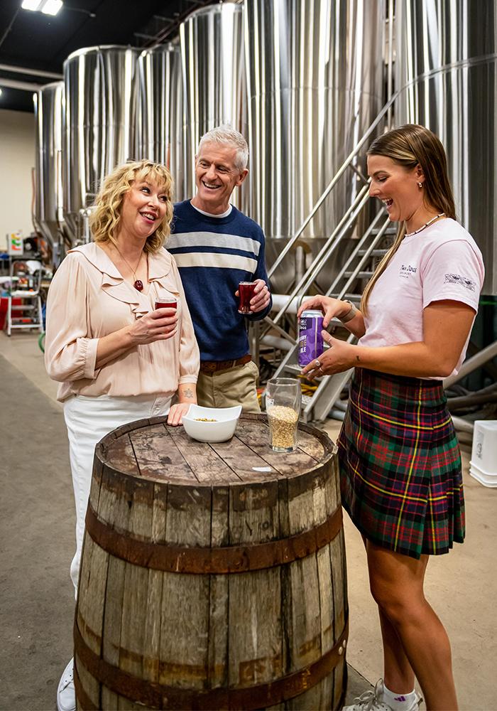 A guide showcases a can of Twa Dogs beer at Macaloney's Island Distillery to a couple on a tasting tour in Victoria, BC