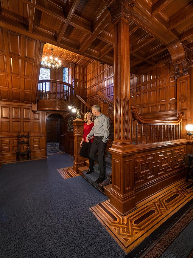 A couple walks hand-in-hand down a wooden staircase inside of Craigdarroch Castle, a National Historic Site in Victoria, BC