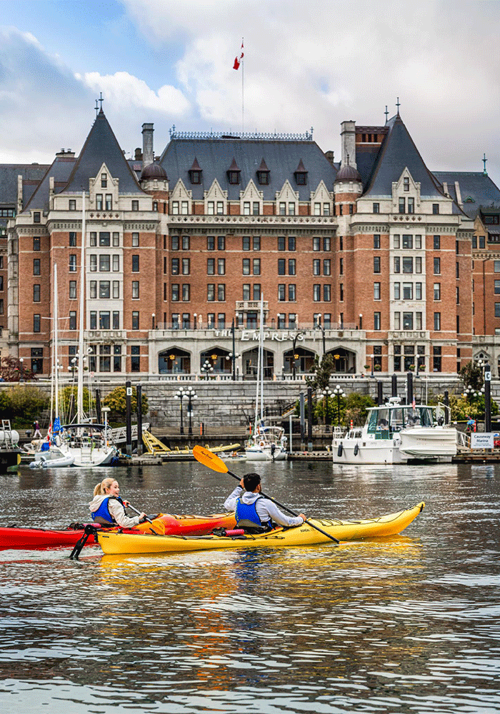 A couple in yellow and red kayaks paddle across the Inner Harbour past the Fairmont Empress hotel in Victoria, BC