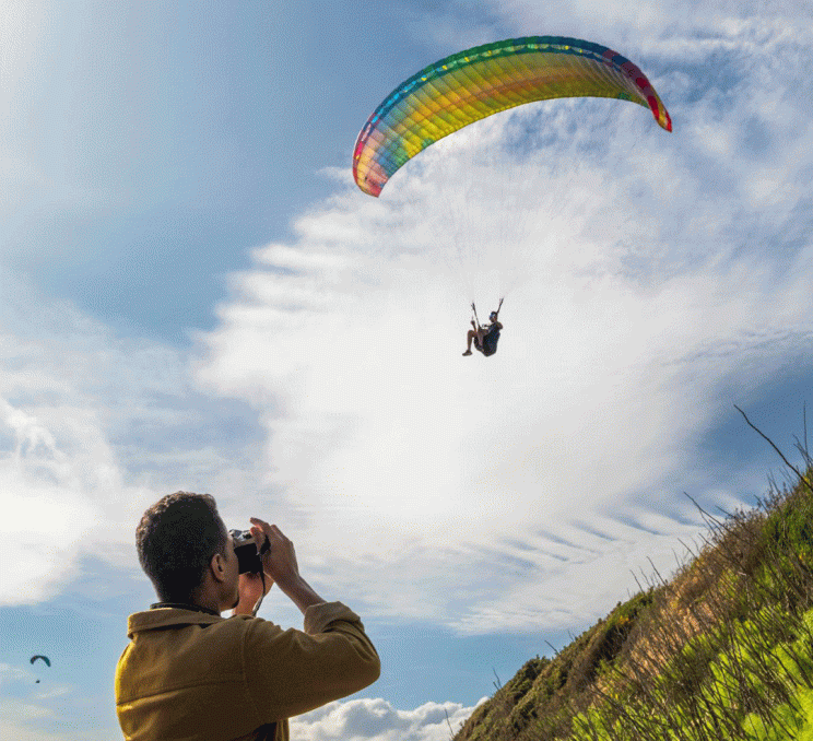 A person takes a photo of a paraglider sailing above Dallas Road in Victoria, BC