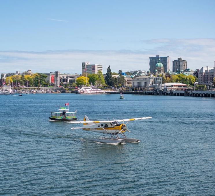 A Kenmore Air seaplane taxis across Victoria, BC's Inner Harbour while harbour ferries pass in behind