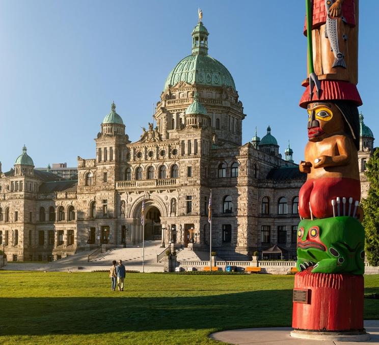 A couple walks on the lawn of the BC Parliament Buildings in Victoria, BC at sunrise. There is a Indigenous carving in the foreground.
