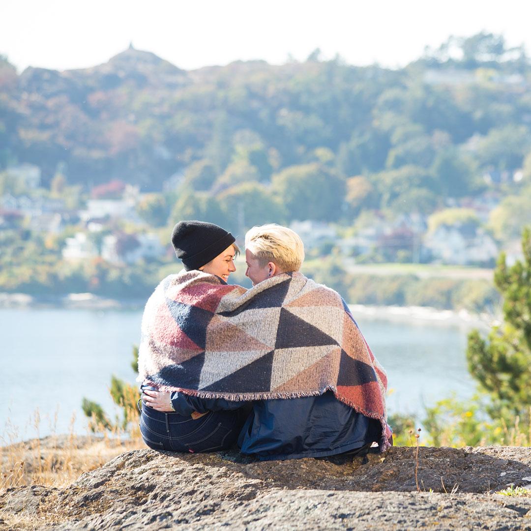 A female couple wrapped in a blanket enjoys a date in a park overlooking the ocean in Victoria, BC