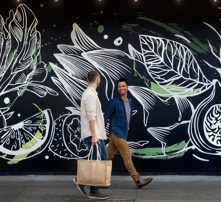 A male couple carrying reusable bags walks past the Root Cellar Village Green Grocer in Cook Street Village, part of Victoria, BC's Fairfield neighbourhood