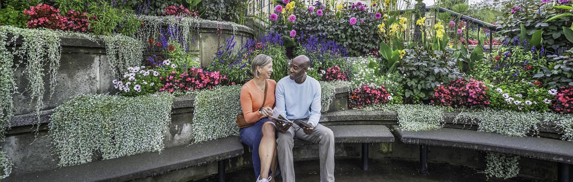 A couple reads a copy of the Victoria Vacation Guide while sitting on a bench at the Inner Harbour outside the Victoria Visitor Centre while surrounded by flowers in Victoria, BC