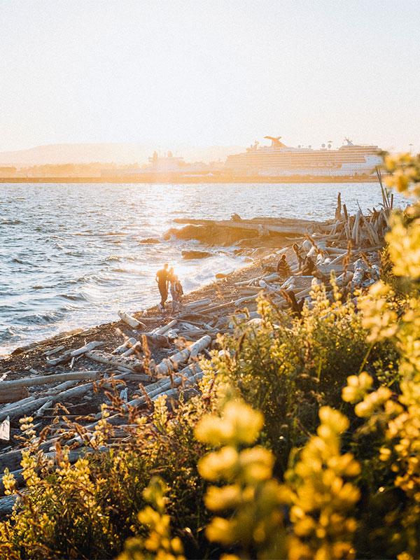 A view from the Dallas Road pathway at sunset in Victoria, BC shows a couple walking the beach and a cruise ship in the distance