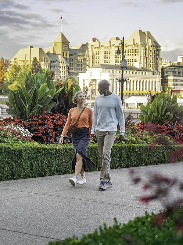 A couple walks hand-in-hand along a walkway in front of the Fairmont Empress on Victoria, BC's Inner Harbour