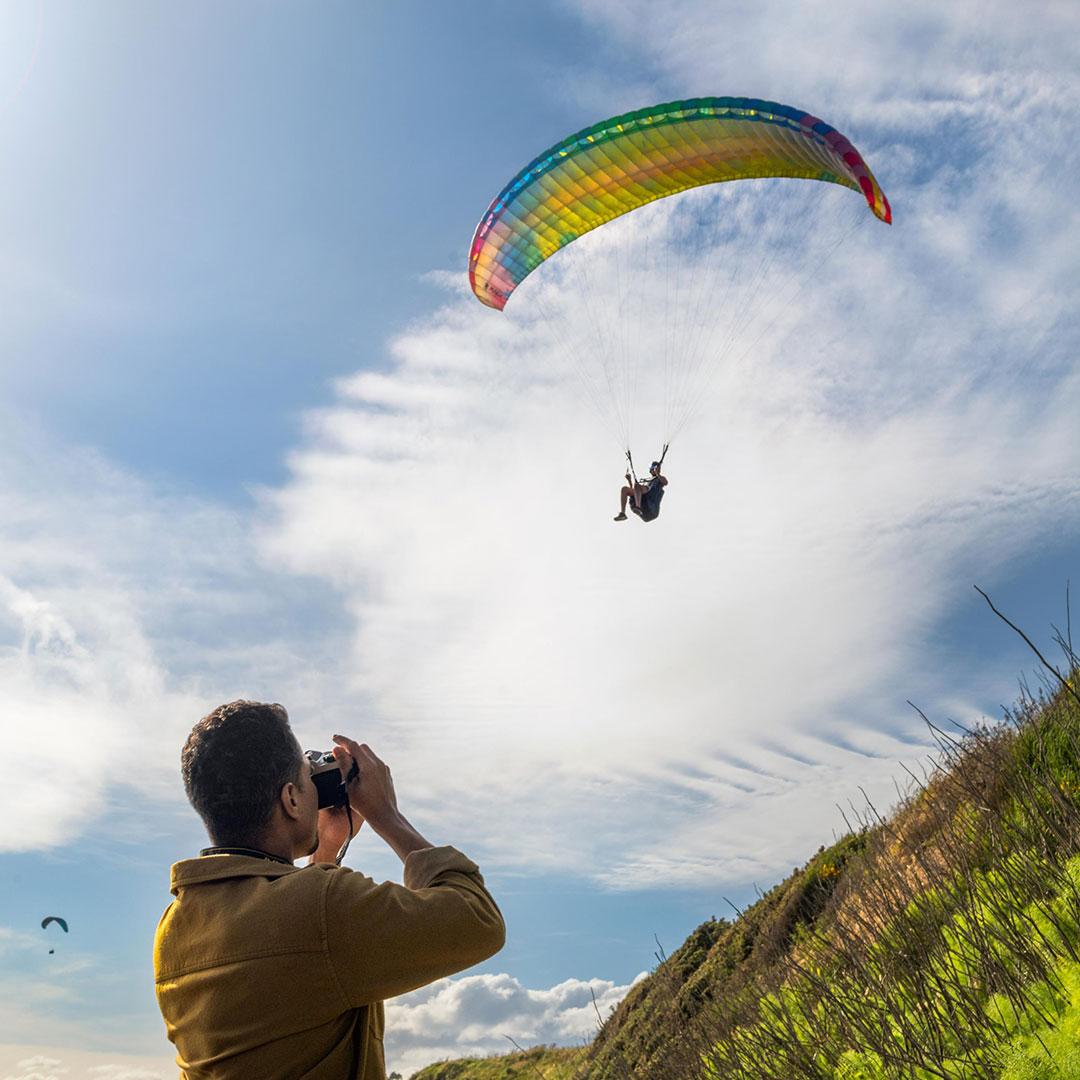A person takes a photo of a paraglider flying overhead at Clover Point along the Dallas Road water font in Victoria, BC