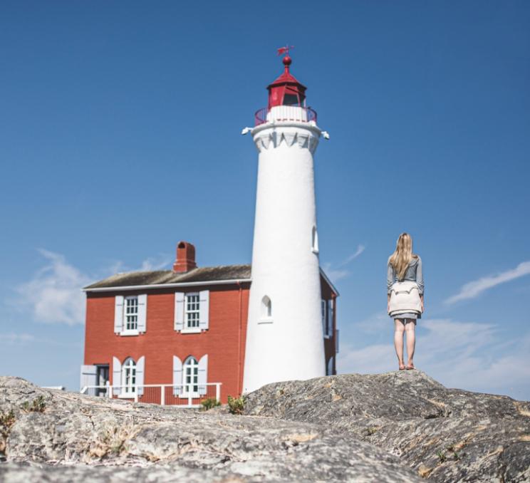 A visitor stands on rocky shoreline beside Fisgard Lighthouse at Fort Rodd Hill National Historic Site in Colwood, overlooking the coast on a clear day.