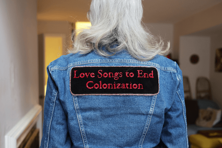 Person wearing a denim jacket with an embroidered patch reading "Love Songs to End Colonization" on the back.