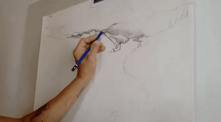 Close-up of an artist sketching a detailed landscape scene with a blue pencil on a large white canvas.