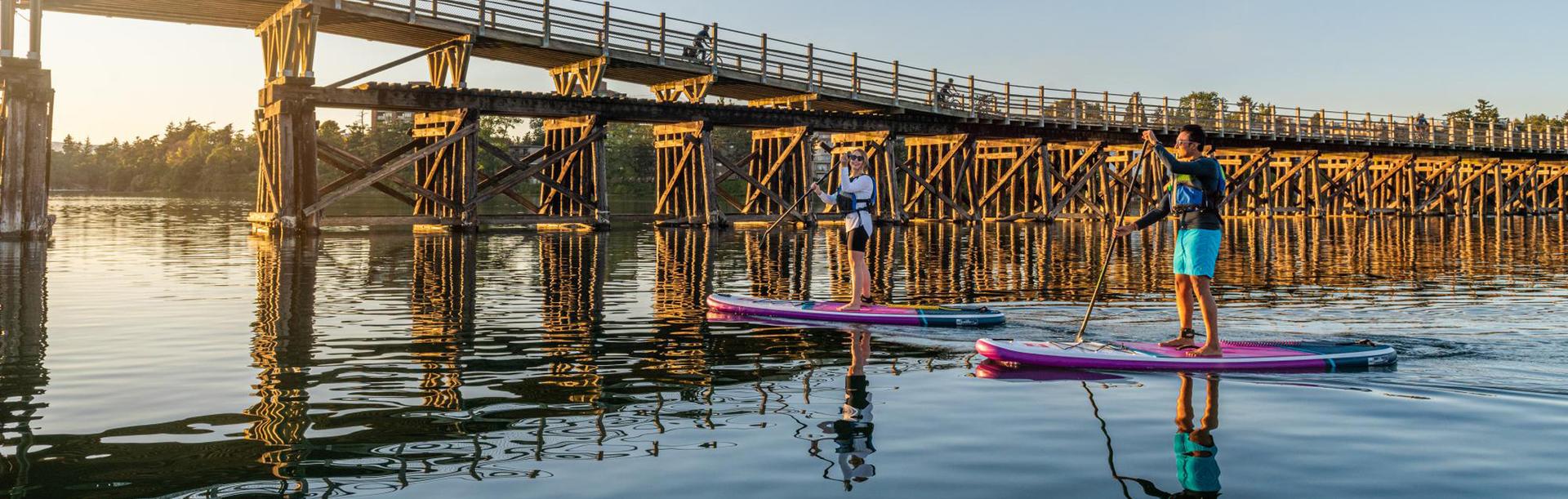 Two people paddleboard beneath the historic Selkirk Trestle spanning the Gorge Waterway in Victoria during a golden summer sunset.