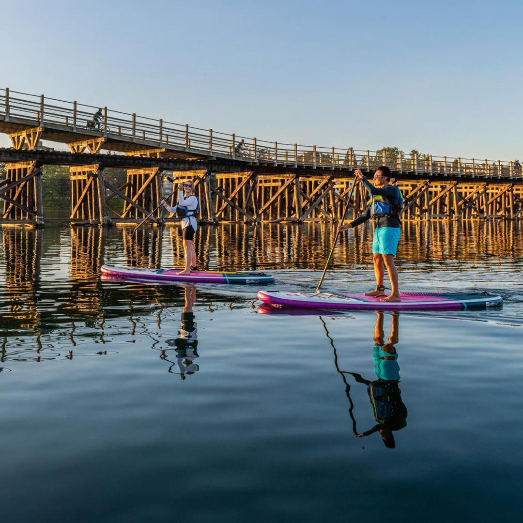 Two people paddleboard beneath the historic Selkirk Trestle spanning the Gorge Waterway in Victoria during a golden summer sunset.