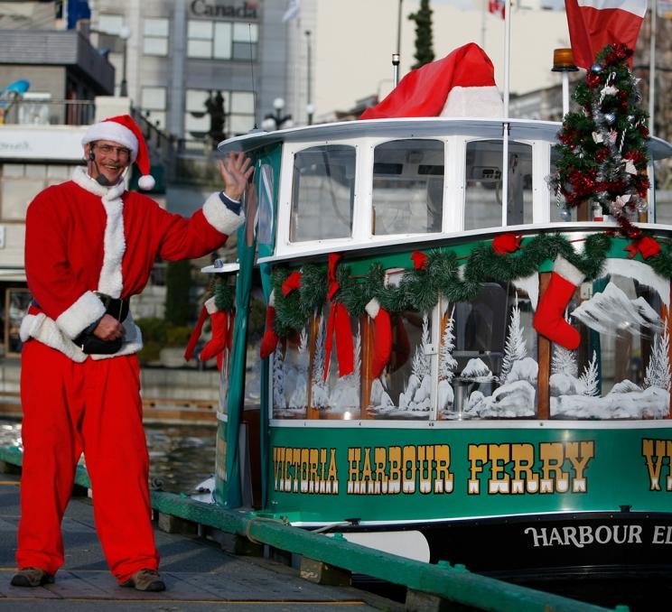 A Victoria Harbour Ferry Captain, dressed as Santa Claus, stands beside his vessel, which is adorned in garland on Victoria, BC's Inner Harbour