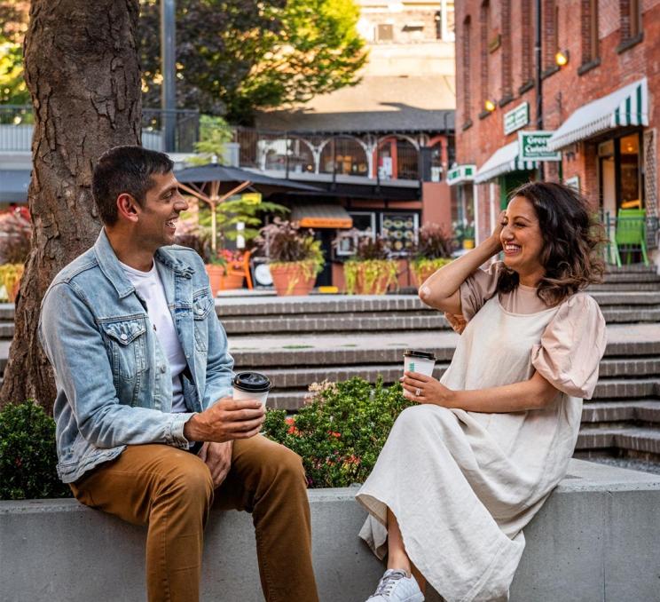 A man in a denim jacket and a woman in a white dress enjoy coffee in Market Square in Victoria, BC