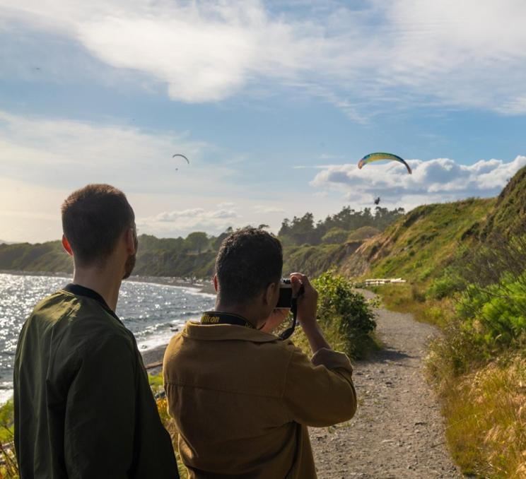 A couple walking along the beachfront of Dallas Road takes a photo of a paraglider overhead in Victoria, BC