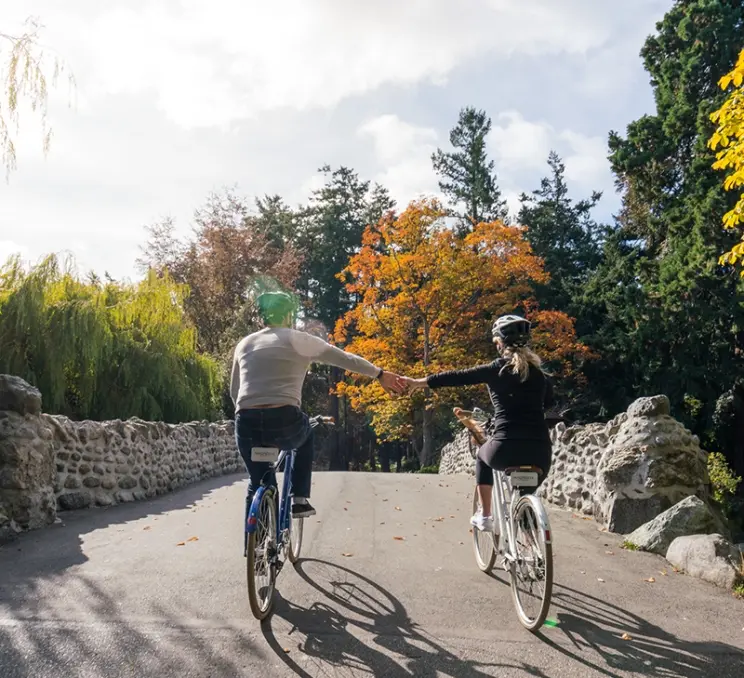 A couple rides bikes, hand-in-hand over the stone bridge in Beacon Hill Park in Victoria, BC