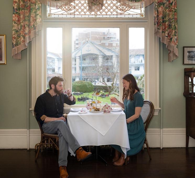 A woman in a teal dress and a man in a black dress shirt enjoy afternoon tea, window-side at the Pendray Teahouse in Victoria, BC