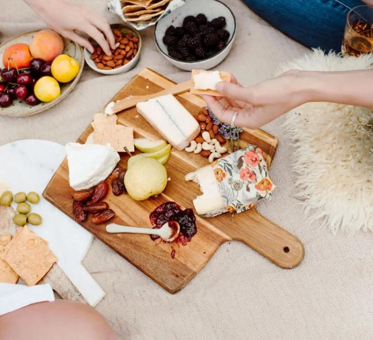 A picnic on a picnic blanket shows a charcuterie board that features a piece of cheese wrapped in Goldlilocks Good's beeswax wrap, in Victoria, BC