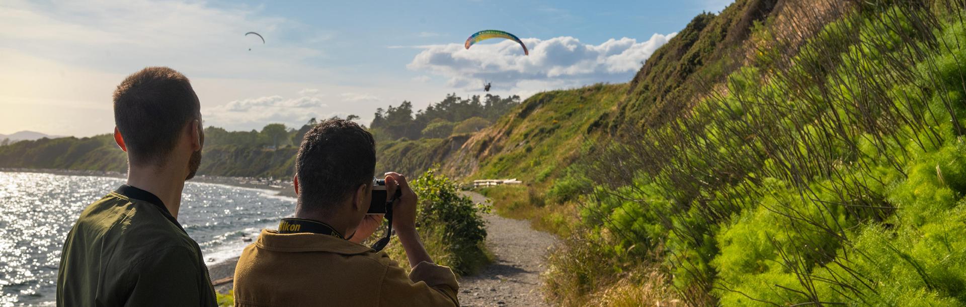 A photographer takes a photo of a paraglider flying over the beach at Clover Point in Victoria, BC