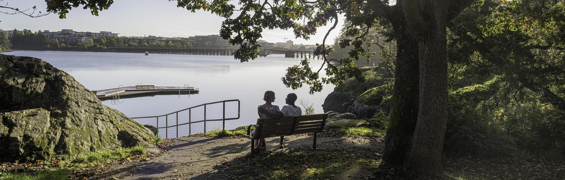 A couple sat on a bench in Banfield Park looks out over the Gorge Waterway in Victoria, BC