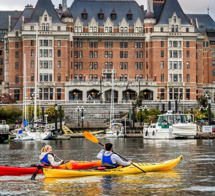 A young couple in red and yellow kayaks paddle across the Inner Harbour in front of the Fairmont Empress Hotel in Victoria, BC
