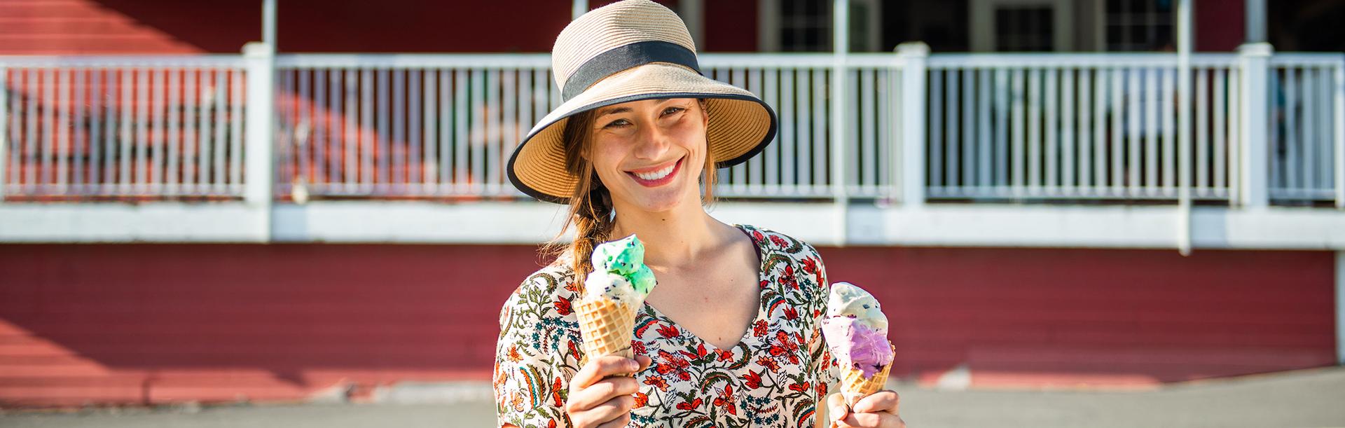 A woman in a sunhat stands outside of Red Barn Market holding two ice cream cones in Victoria, BC