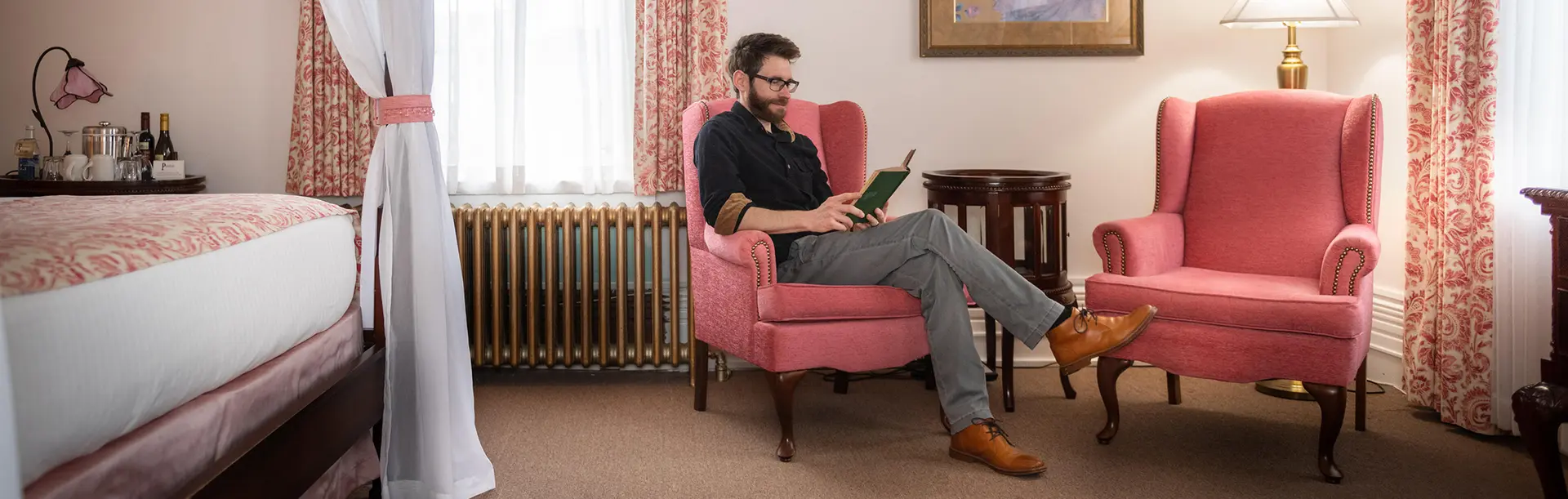 A man reads a book while sat on a pink seat in a hotel room in Victoria, BC