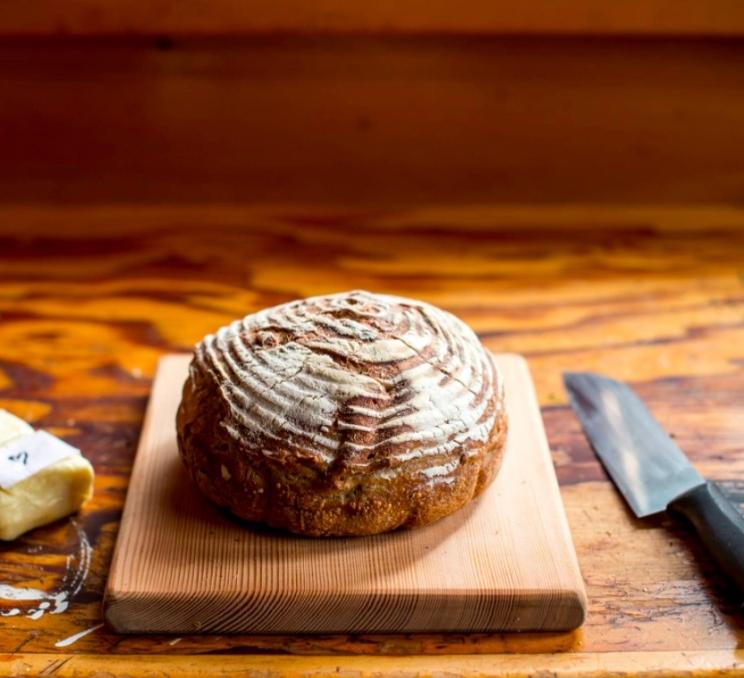 A loaf of gluten free board on a cutting board, with a bread knife on the right and a block of cheese on the left.