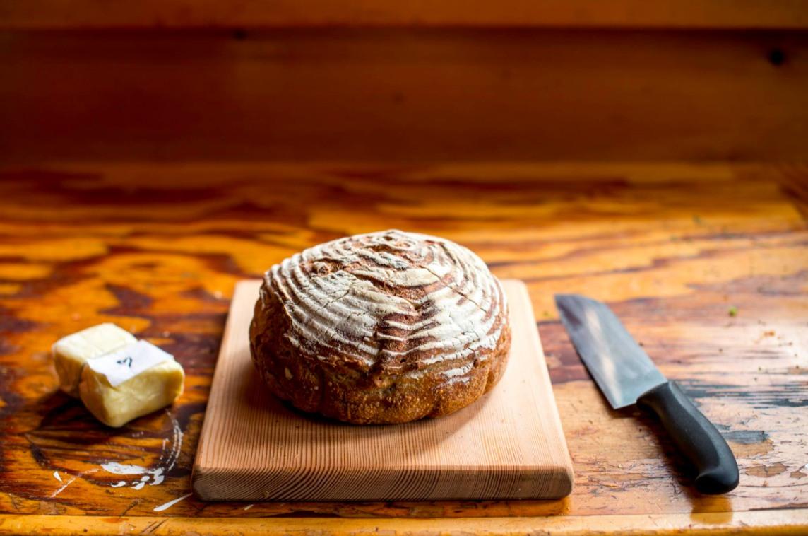 A loaf of gluten free board on a cutting board, with a bread knife on the right and a block of cheese on the left.
